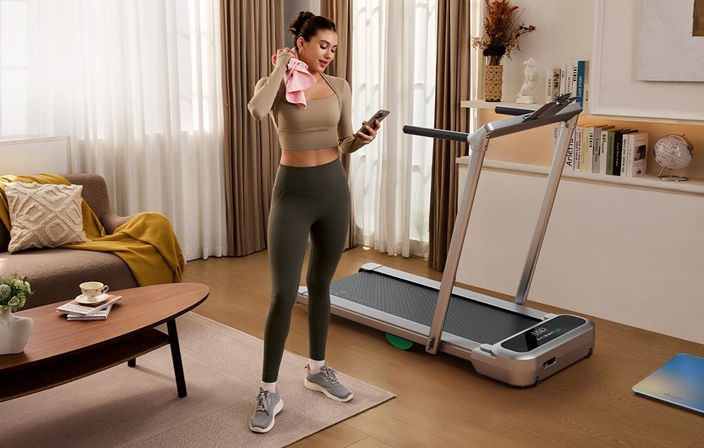 Žena v športovom oblečení s uterákom a telefónom stojaca vedľa zloženého bežeckého pásu v obývacej izbe