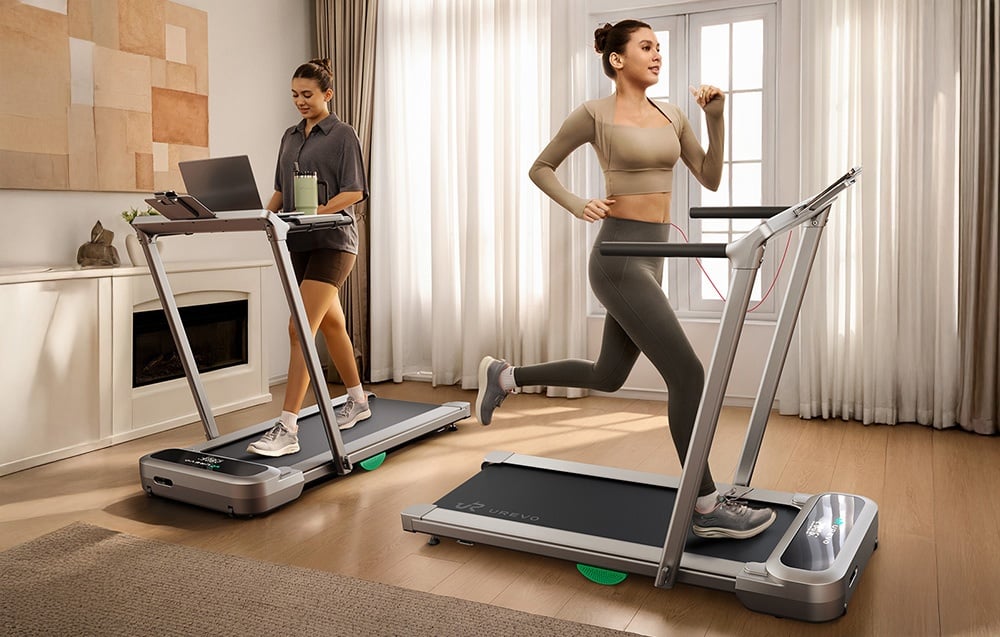 Dve ženy v obývacej izbe, jedna pracuje za stolom na bežeckom páse, druhá beží na stacionárnom bežeckom páse