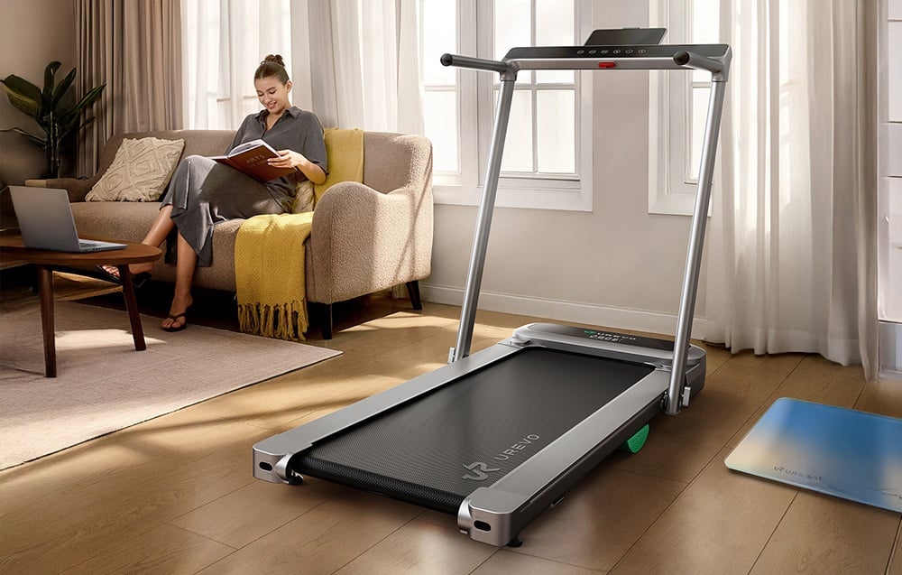Stacionárny bežecký pás stojaci v obývačke, v pozadí žena čítajúca knihu na pohovke
