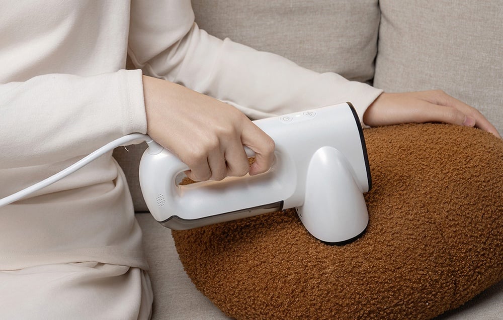 Osoba v béžovom oblečení sedí na pohovke a bielym naparovačom čistí hnedý vankúš