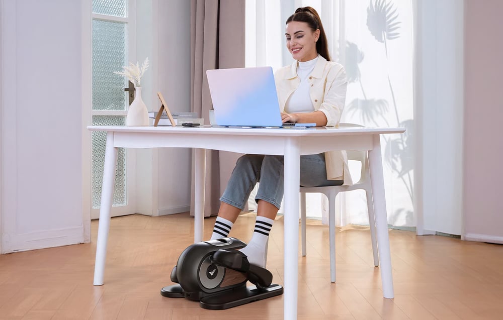 Žena pracuje na svojom notebooku pri stole, pričom pod nohami používa mini eliptický trenažér Merach.