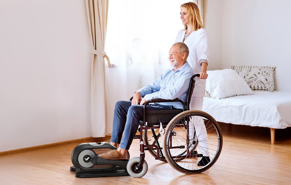 Starší muž cvičí na mini eliptickom trenažéri Merach, zatiaľ čo sedí na invalidnom vozíku, v sprievode svojho opatrovateľa.