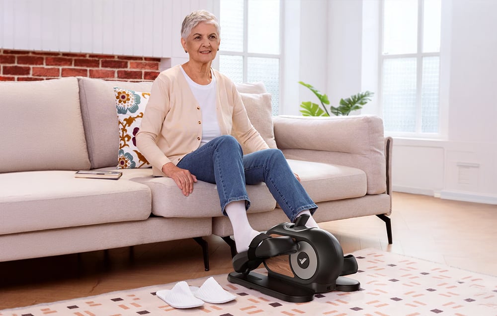 Staršia žena sedí na pohovke a cvičí na mini eliptickom trenažéri Merach vo svojej obývacej izbe.