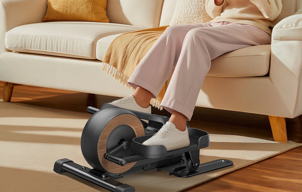 Záber na nohy cvičiacich na mini eliptickom trenažéri Merach umiestnenom vedľa pohovky.