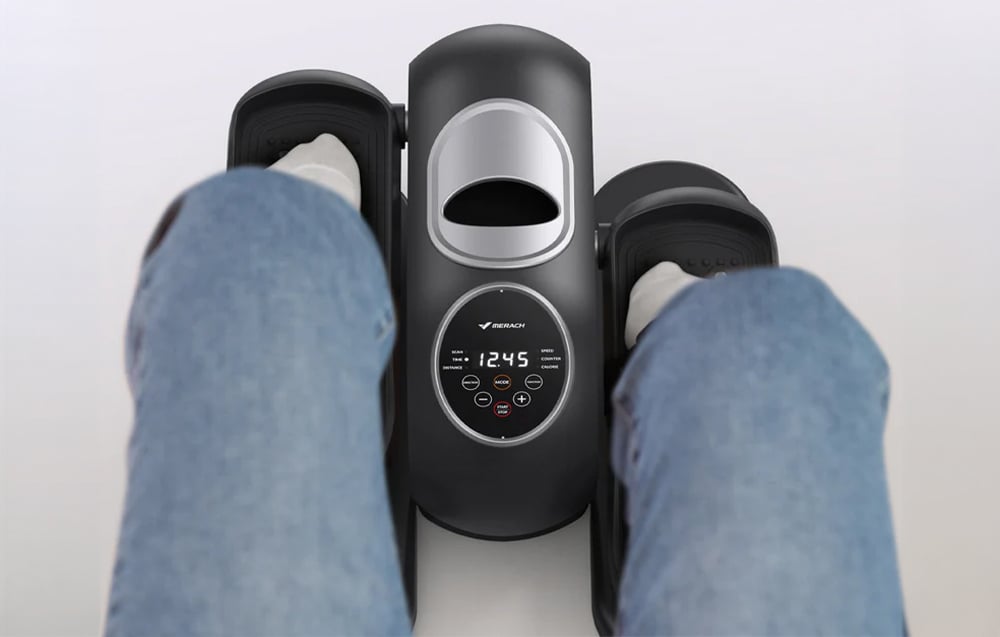 Detailný pohľad na mini eliptický trenažér Merach používaný pod stolom, pohľad zhora na nohy a ovládací panel s displejom.