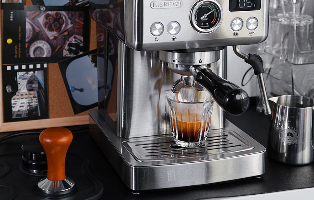 Espresso stroj HiBREW v domácej kuchyni vedľa baristických doplnkov, ako je drevená tamper a kovová kanvica na mlieko.