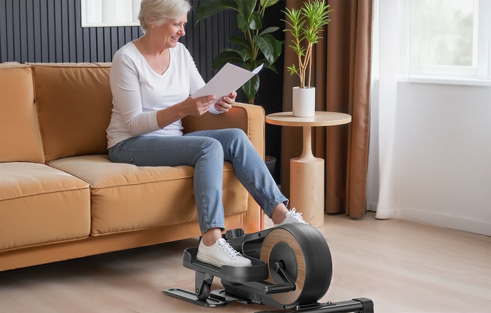 Staršia žena sedí na pohovke a cvičí na mini eliptickom trenažéri Merach v svetlej obývačke.