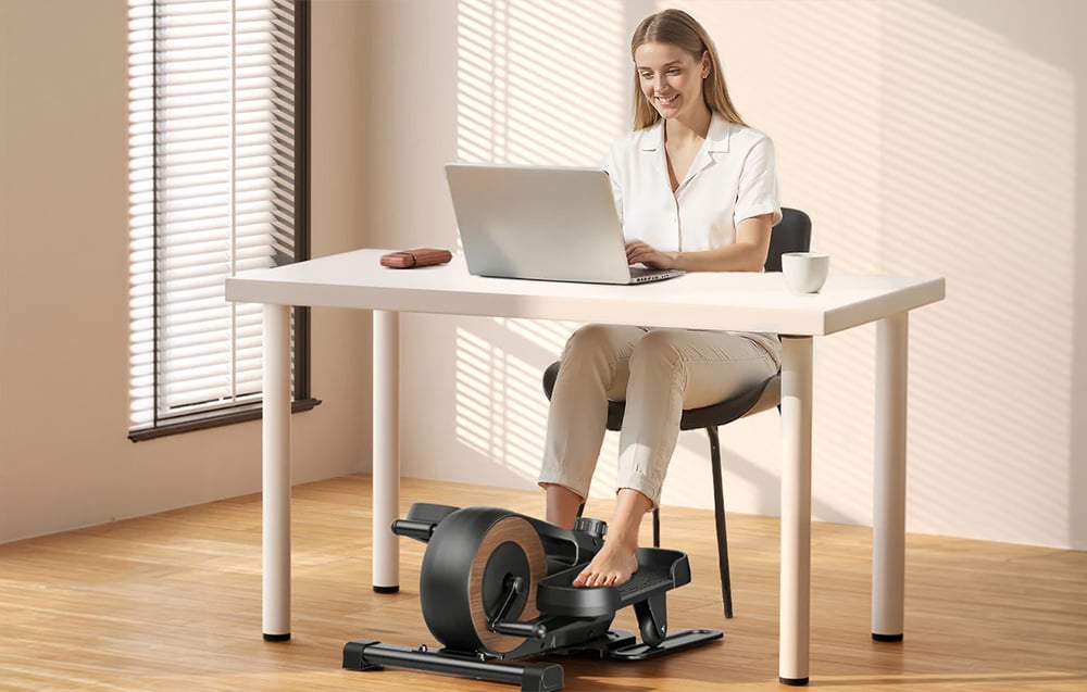 Žena pracuje pri stole na notebooku a zároveň cvičí na mini eliptickom trenažéri Merach pod stolom.