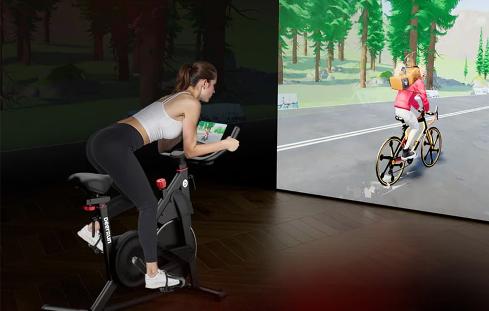 Žena cvičiaca na stacionárnom bicykli počas virtuálnej jazdy zobrazená na veľkej obrazovke