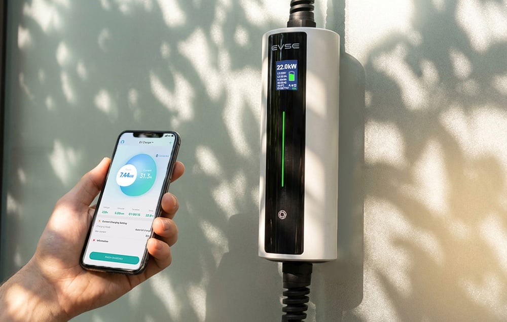 Smartfón v ruke s otvorenou aplikáciou, nabíjačka elektromobilov na stene vedľa neho
