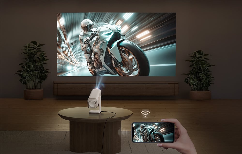 Projektor stojací na stole premieta na stenu dynamickú scénu s motocyklom, pričom obraz je bezdrôtovo prenášaný zo smartfónu.