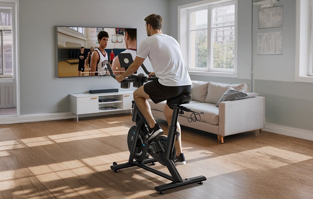 Muž cvičí na stacionárnom bicykli a sleduje zápas v televízii
