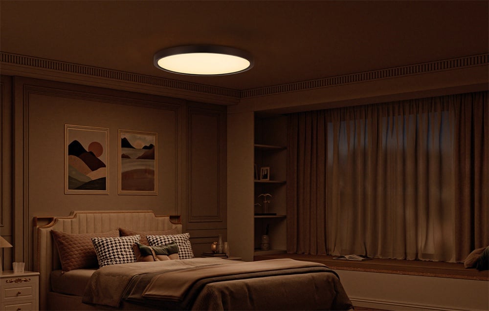 Stropná lampa Yeelight Meteor C230 svietiaca v zatemnenej spálni s béžovou posteľnou bielizňou a obrazmi nad posteľou.