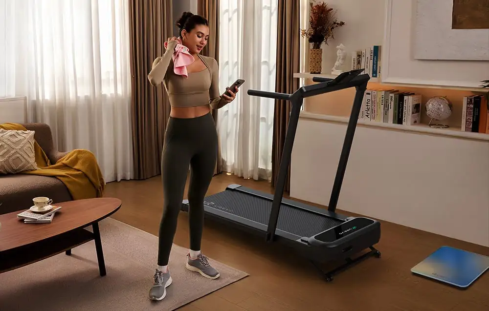 Žena v športovom oblečení stojí vedľa bežeckého pásu, pozerá na telefón, s uterákom na ramene