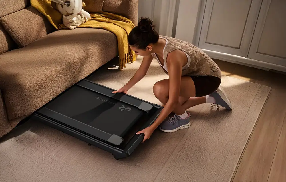 Žena skrývajúca zložený bežecký pás pod pohovkou v obývačke